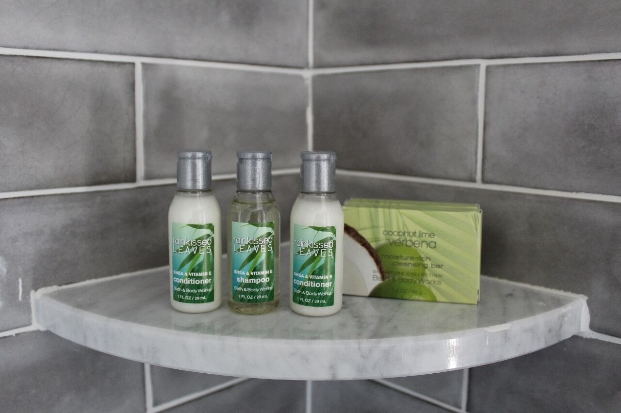 Três garrafas de shampoo e condicionador 'rainkissed leaves' sobre um estante de mármore, em frente a uma parede azulejada.