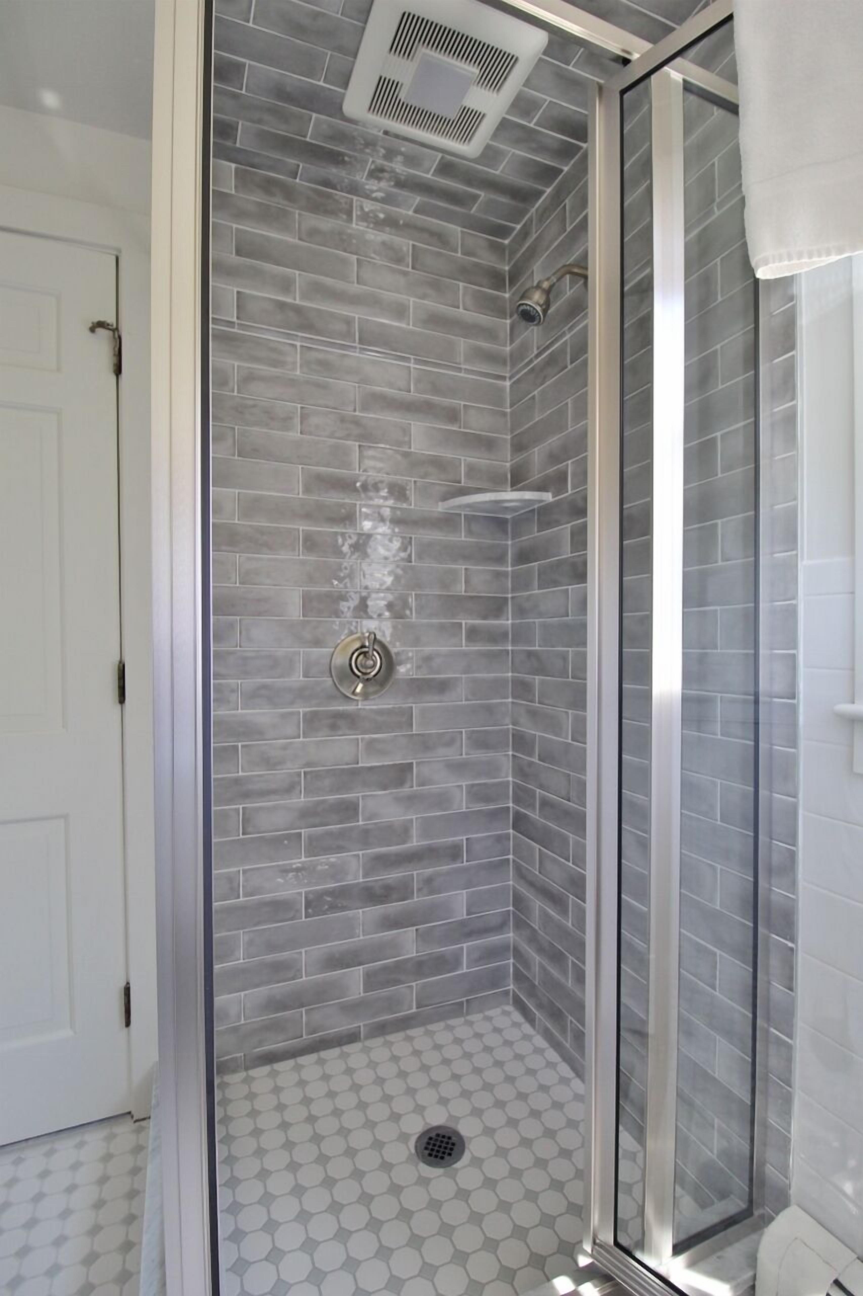 Une douche avec une porte en verre, des murs recouverts de carreaux gris et un sol carrelé de blanc.