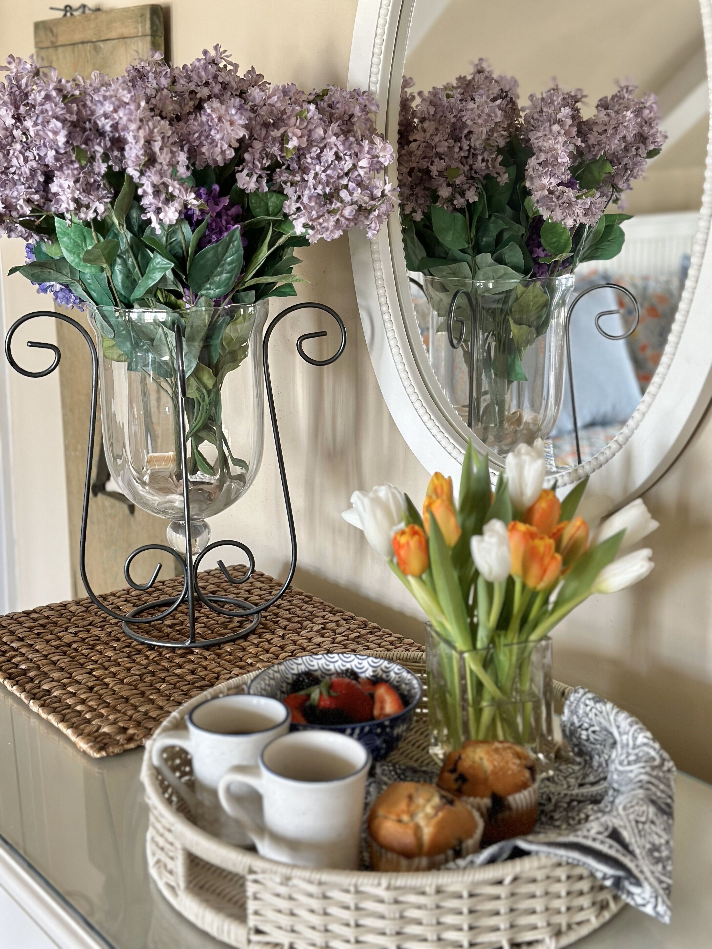 Two clear glass vases with purple flowers on a table, a tray with pastries and a cup, and a mirror reflecting the scene.