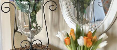 Two clear glass vases with purple flowers on a table, a tray with pastries and a cup, and a mirror reflecting the scene.
