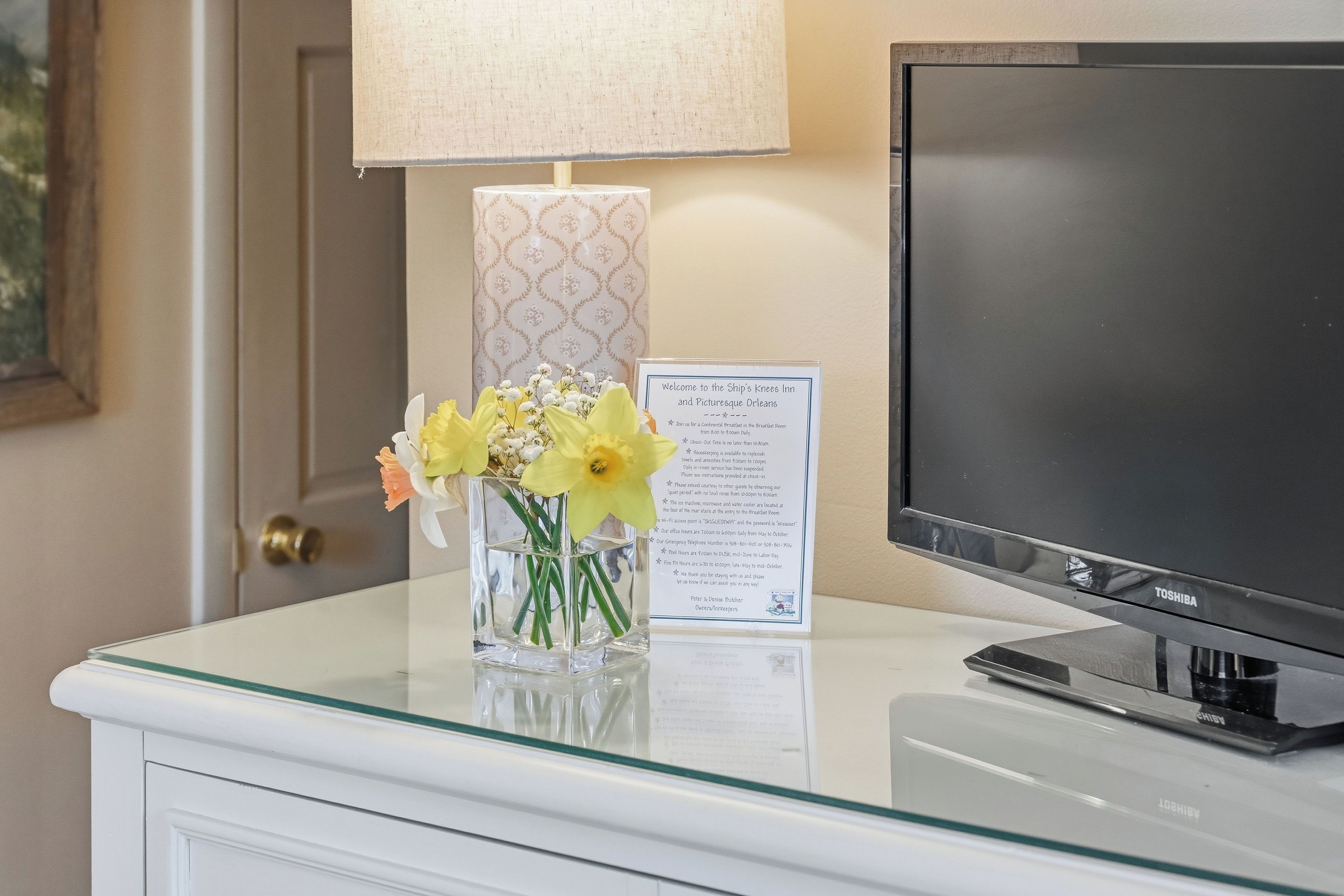 A glass-topped desk with a flat-screen TV, a vase with flowers, a lamp, and a welcome sign.