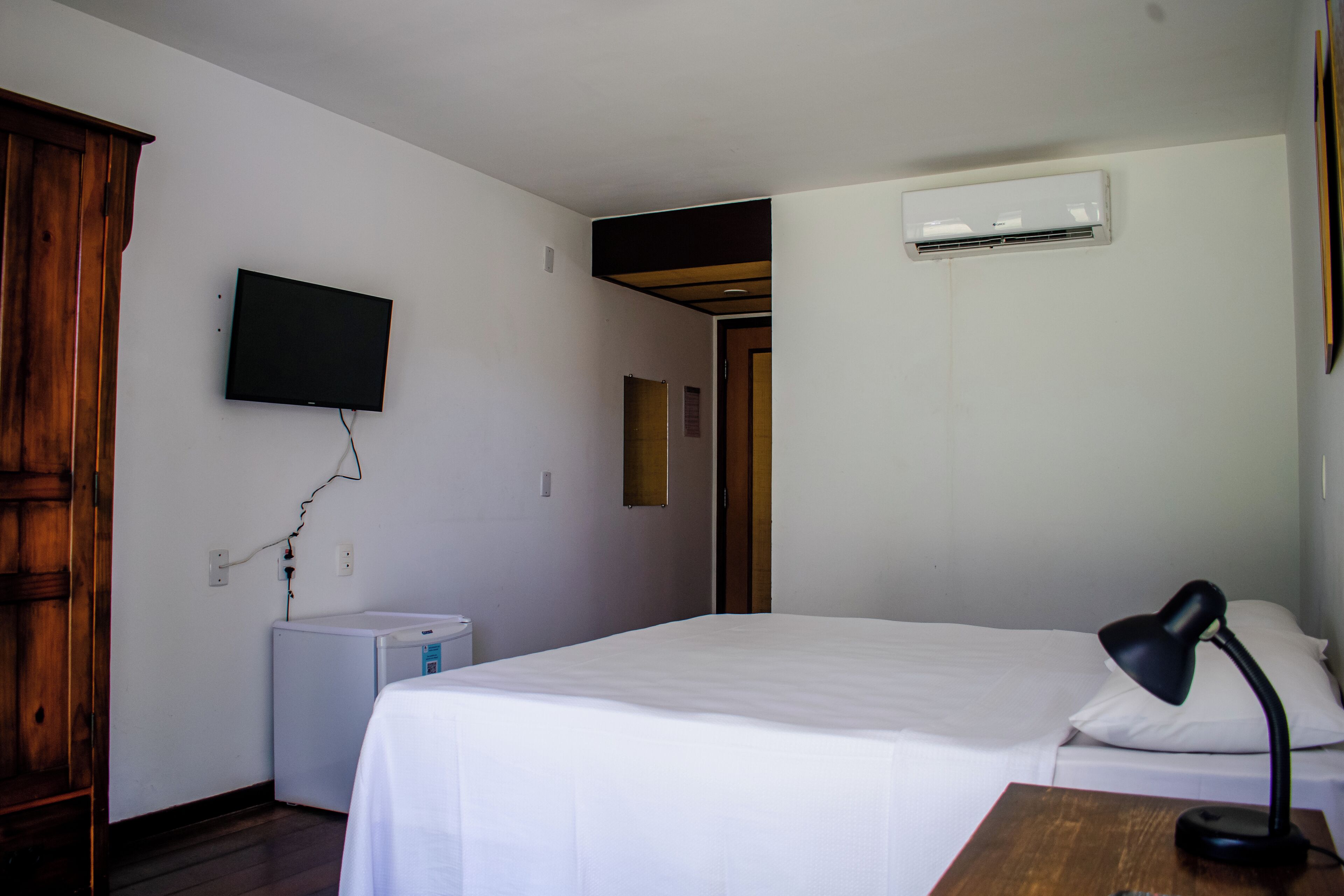 A bedroom with a bed, a desk with a lamp, a television, an air conditioner, and a wardrobe.