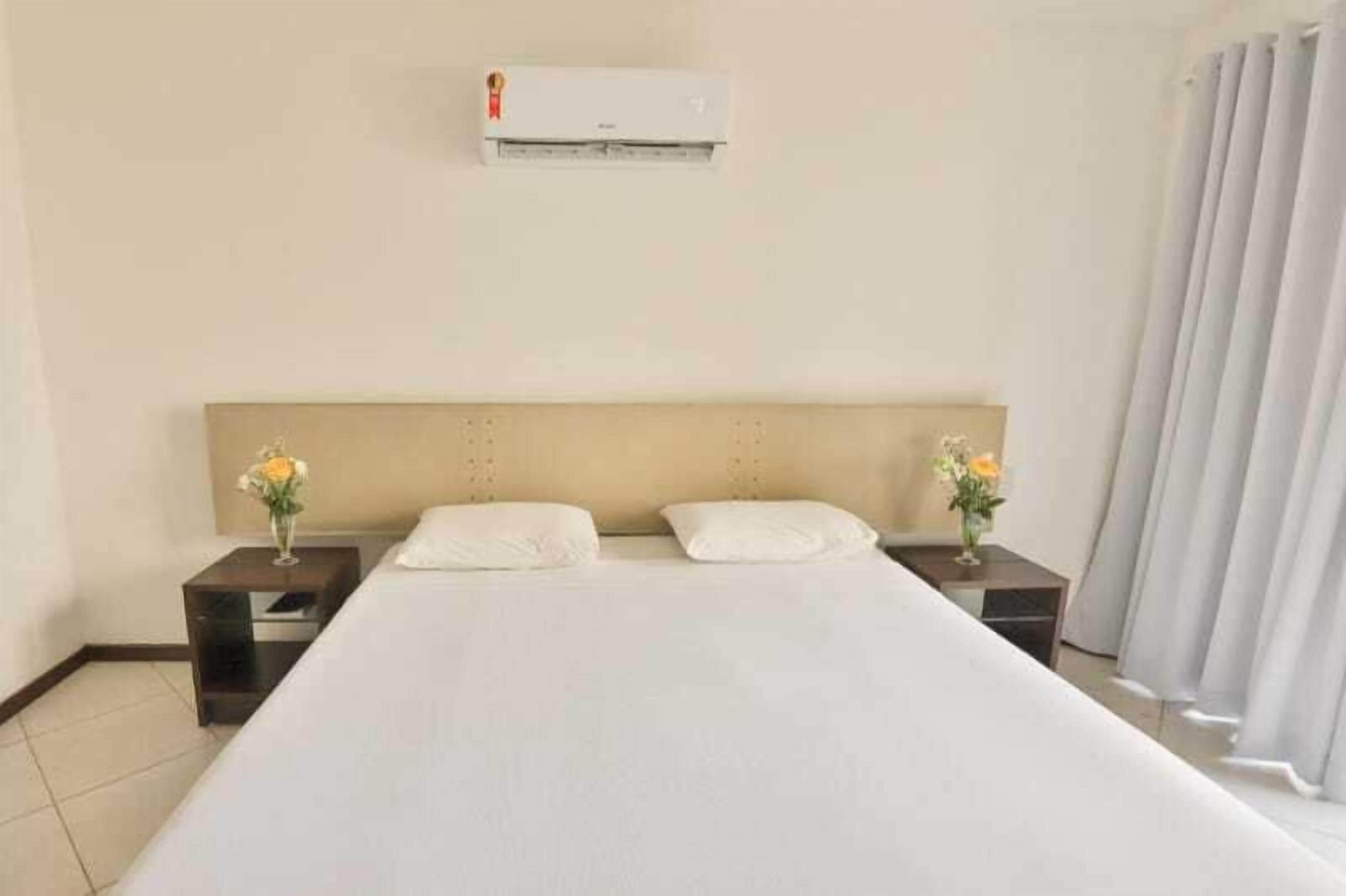 A hotel room with a large bed, two nightstands with flower vases, a wall-mounted air conditioner, and a curtain on the window.