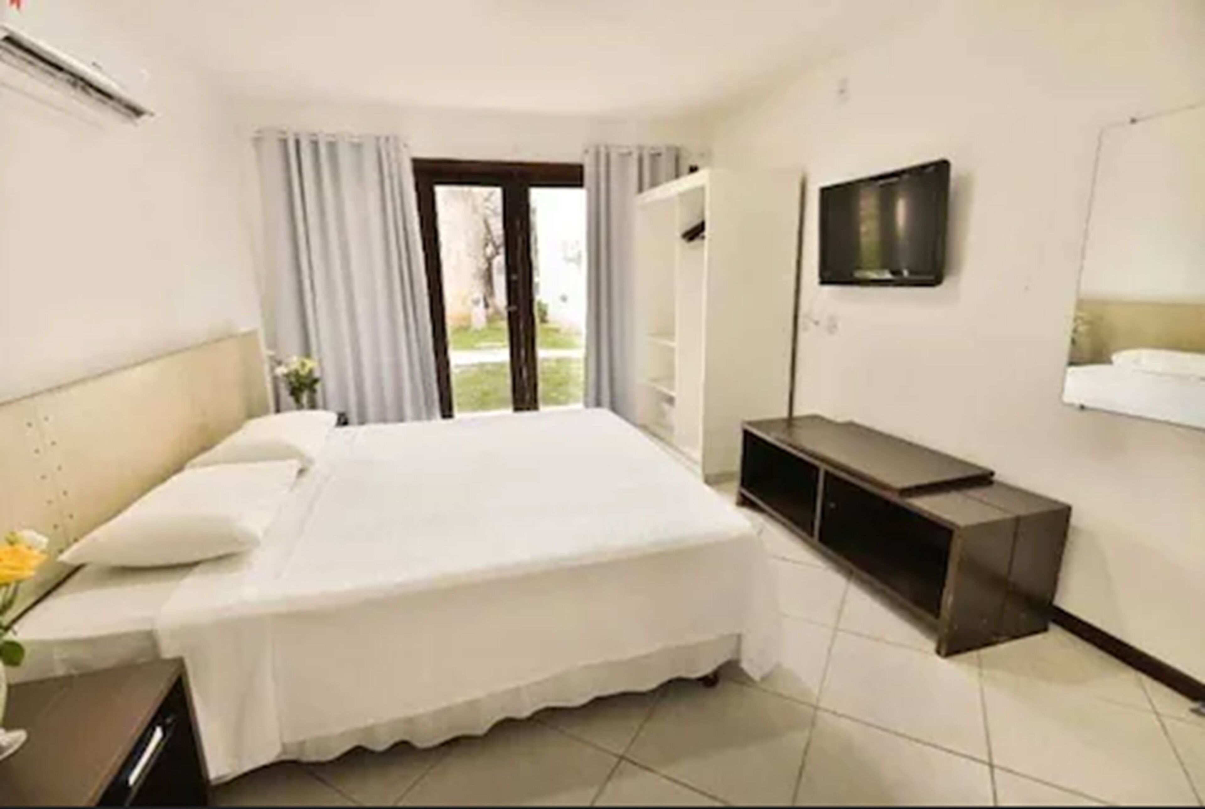 A hotel room with a bed, a TV mounted on the wall, a small desk, and a window with curtains.