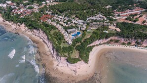 Vista aérea de un complejo turístico en la costa, con playa, piscina y vegetación densa.