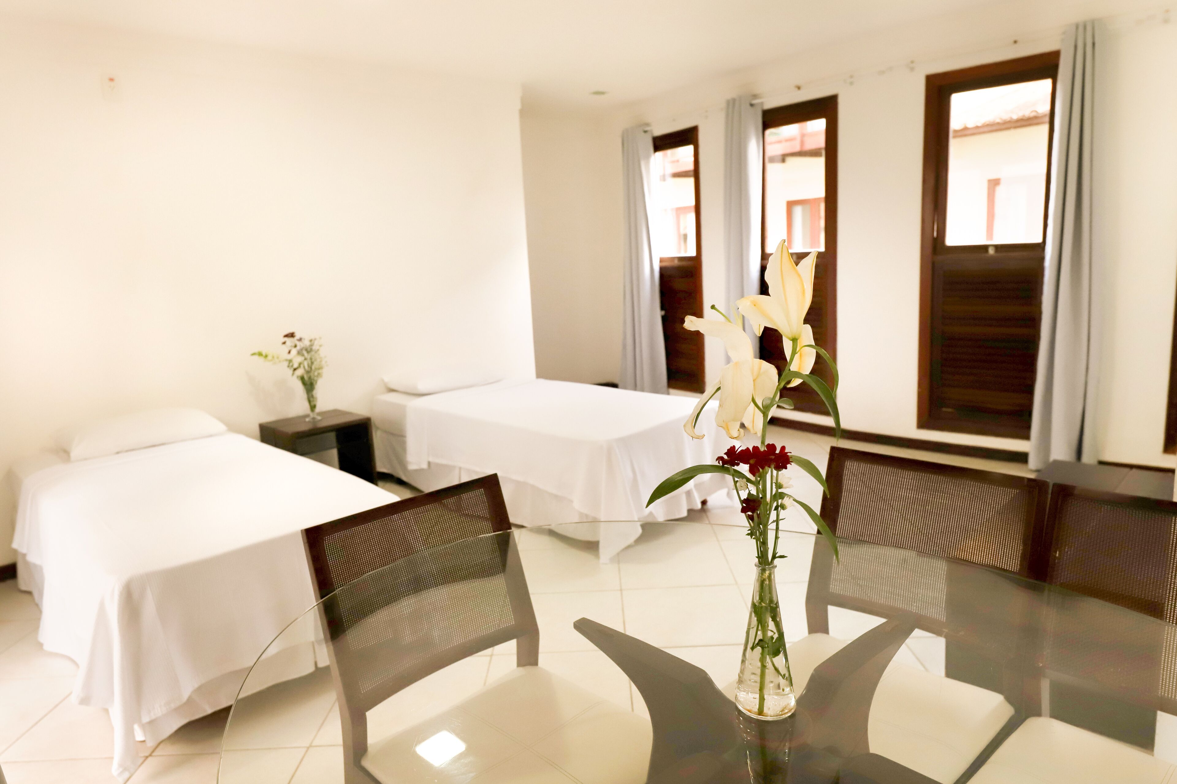A hotel room with two single beds, a dining table with chairs, a vase with flowers, and a window with curtains.