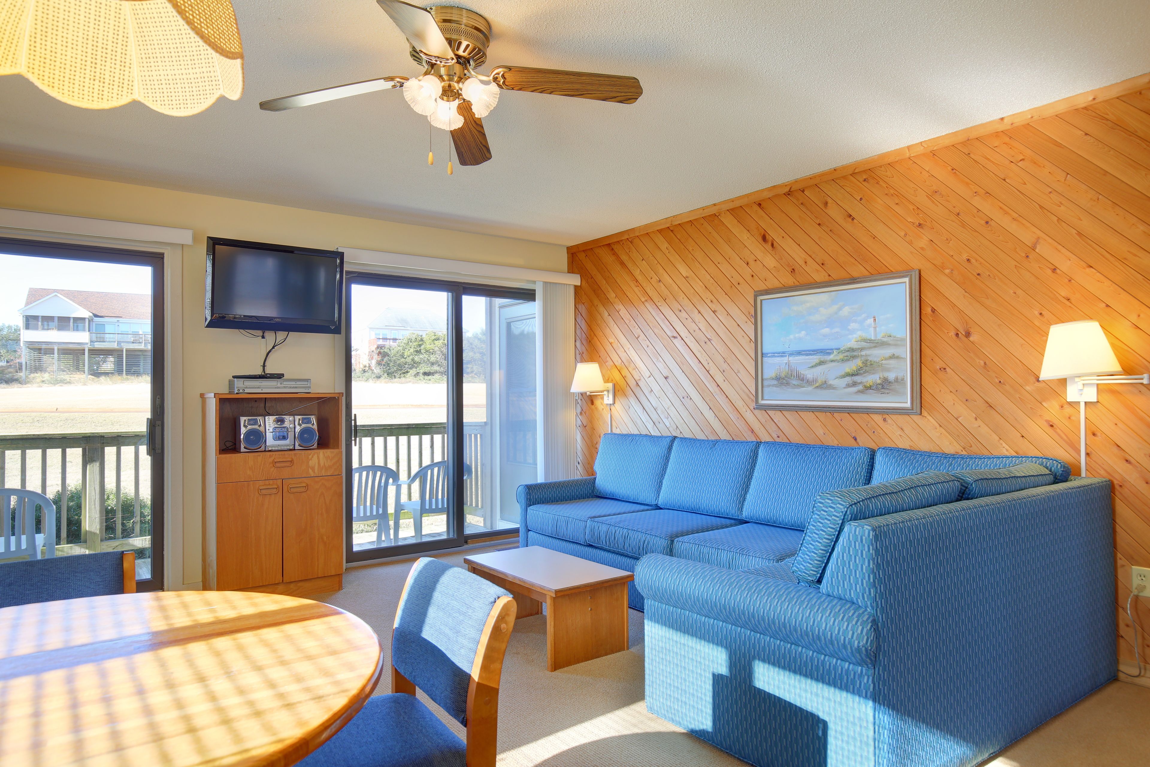 A cozy room with a blue sofa, a wooden table, a ceiling fan, and a TV on the wall.
