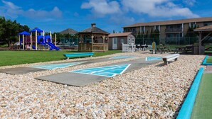 A playground with a slide and climbing structures, a gazebo, picnic tables, and a building in the background. - Sea Scape Beach and Golf Villas (Kitty Hawk)