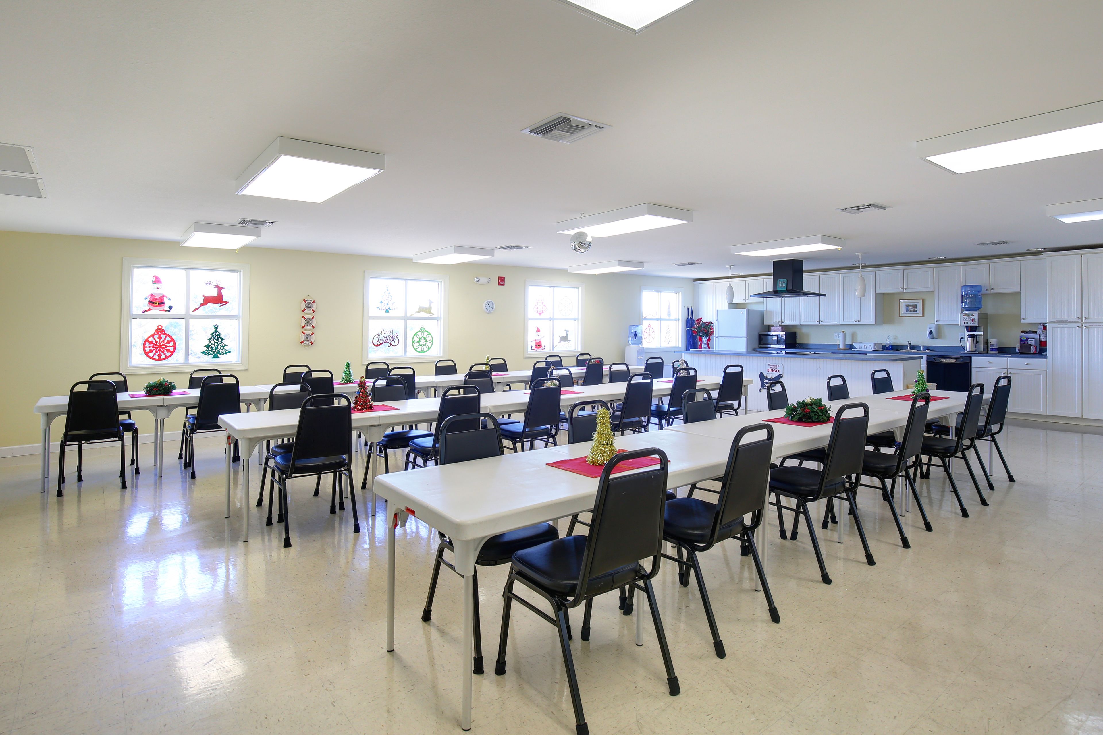 A conference room with multiple tables and chairs, a kitchen area in the background, and holiday decorations on the windows.