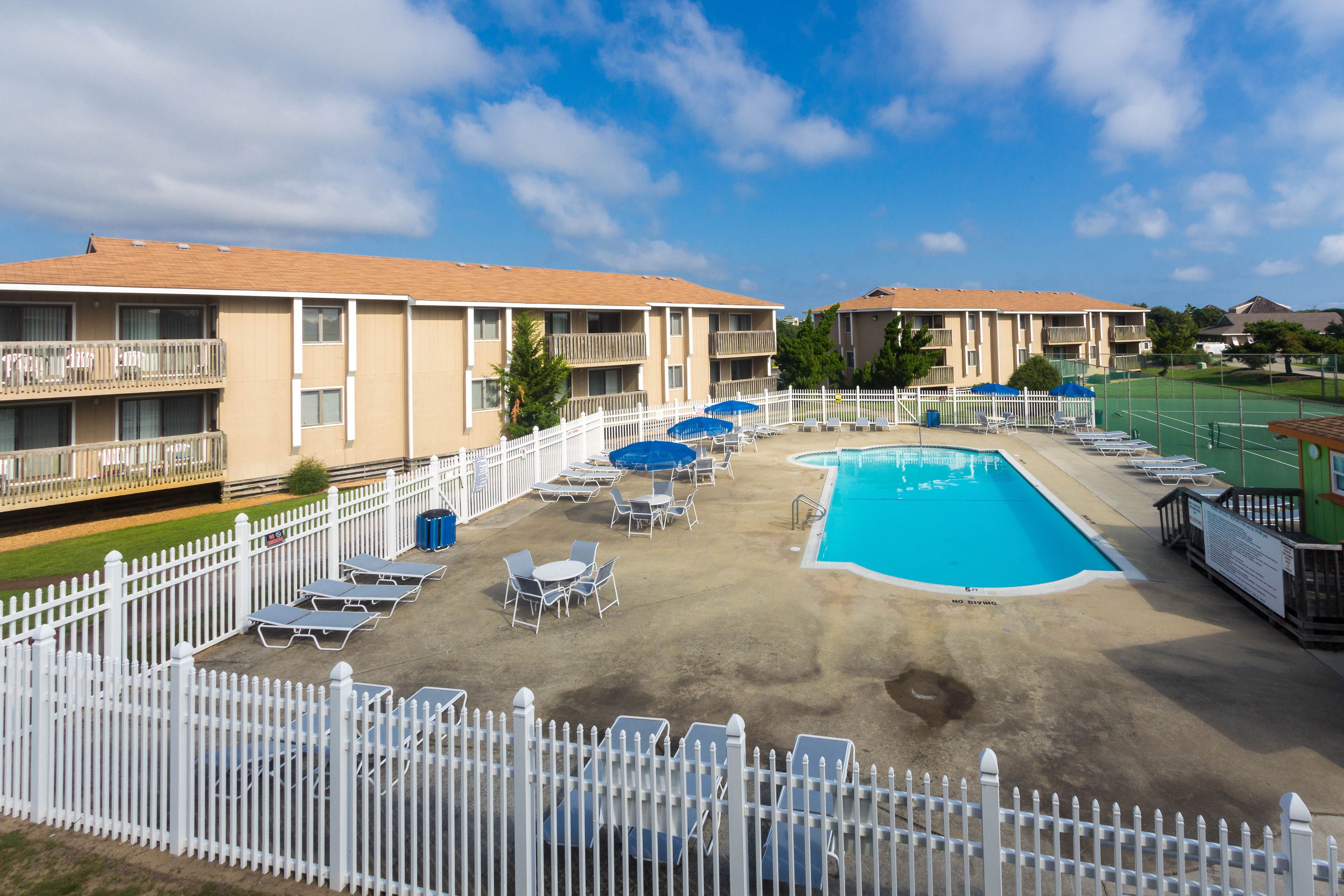 A pool area with lounge chairs and umbrellas, surrounded by a white fence, and adjacent to a building with balconies.