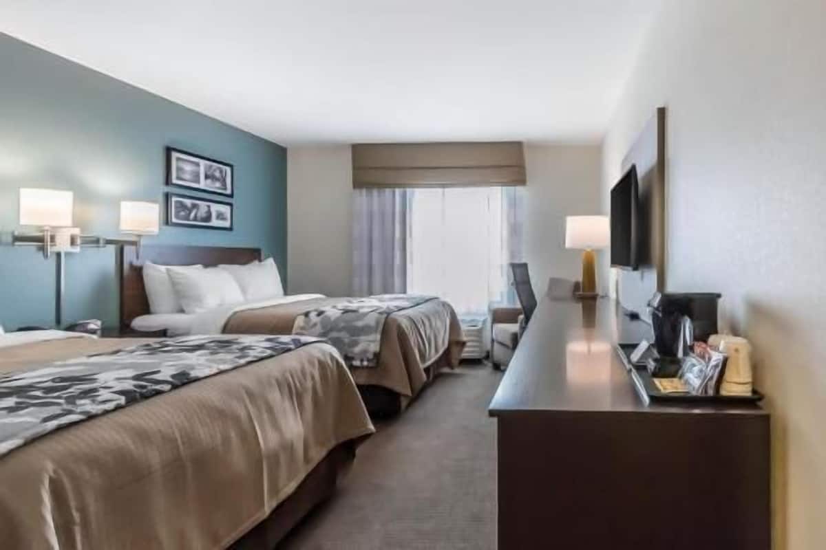 Quarto de hotel com duas camas, uma escrivaninha, televisão e janela com cortinas.
