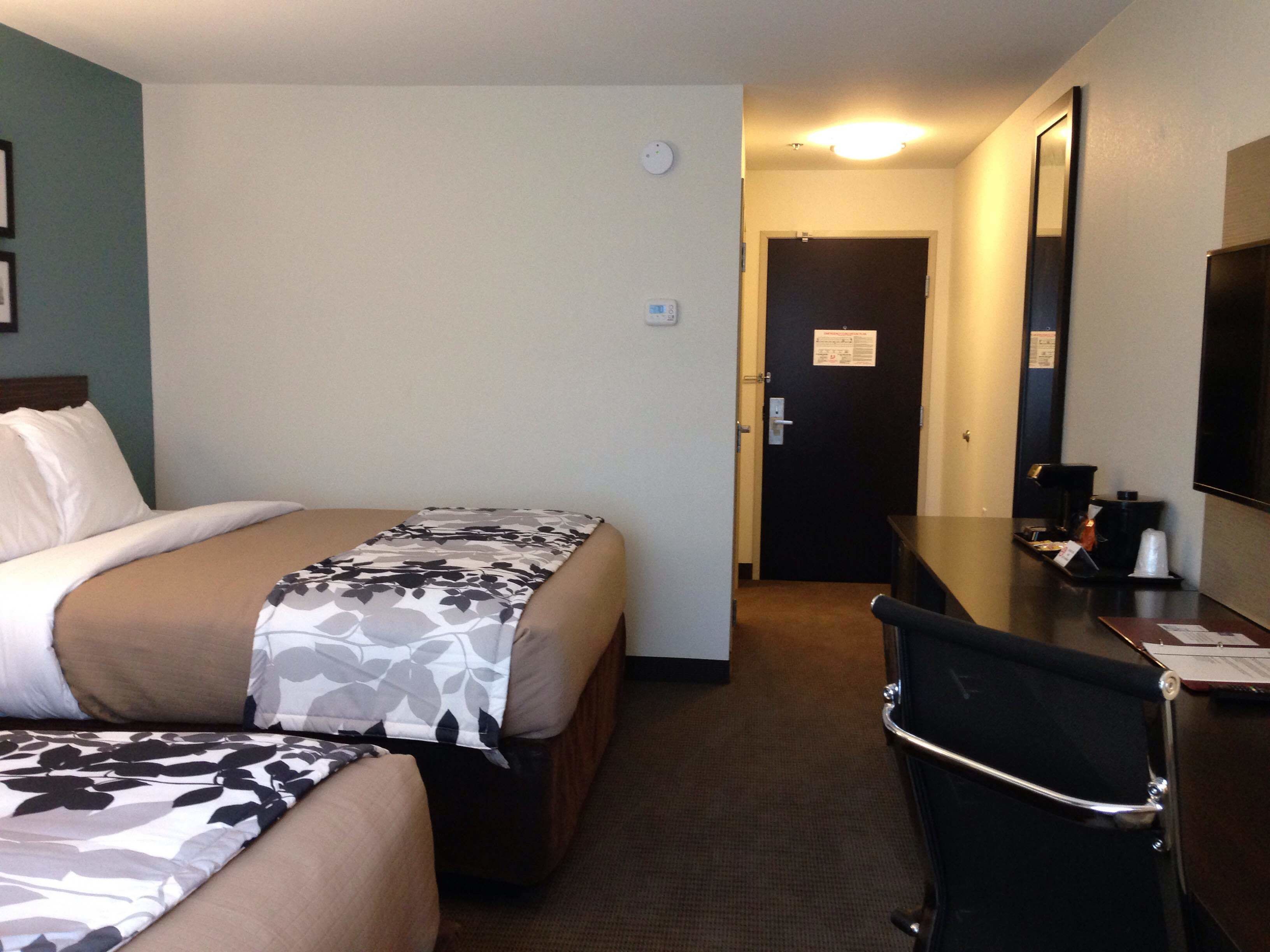 A hotel room with two beds, a desk, a chair, a coffee machine, and a door leading to another room.