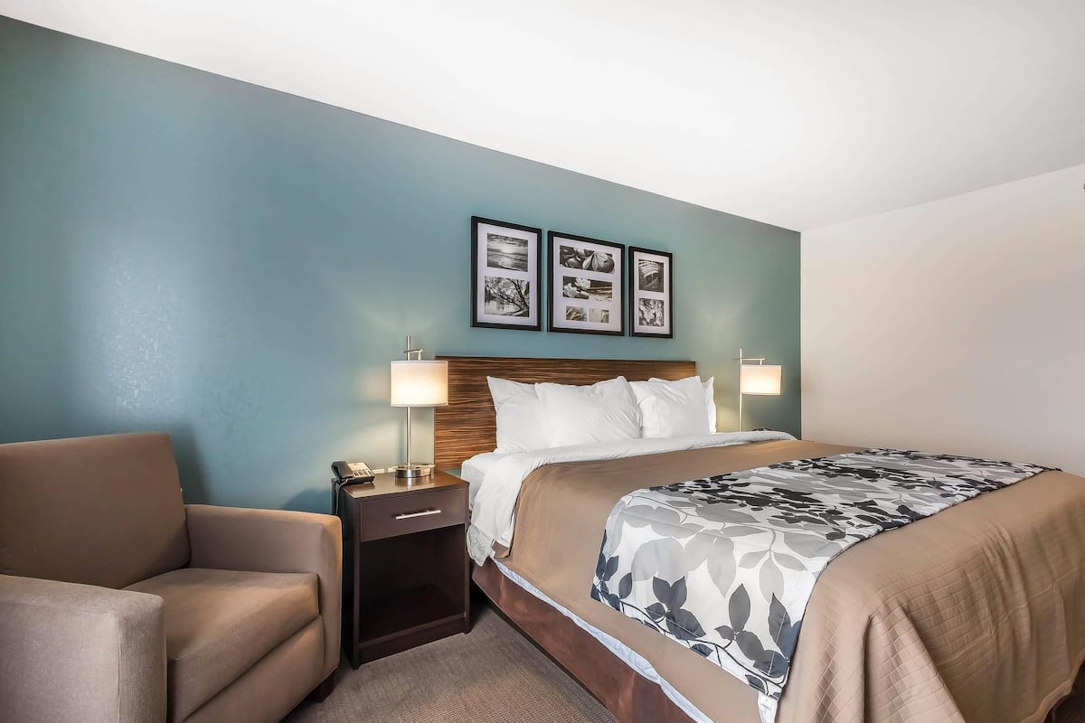 Quarto de hotel com cama, uma cadeira, mesa de cabeceira e quadros na parede.