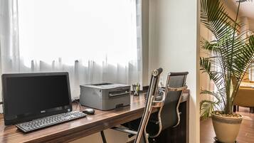 A desk with a Dell laptop, a printer, and a keyboard. There is a chair with a high backrest, a potted plant, and a window with sheer curtains.