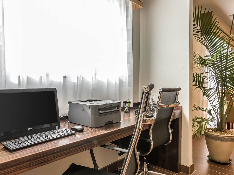 A desk with a Dell laptop, a printer, and a keyboard. There is a chair with a high backrest, a potted plant, and a window with sheer curtains.