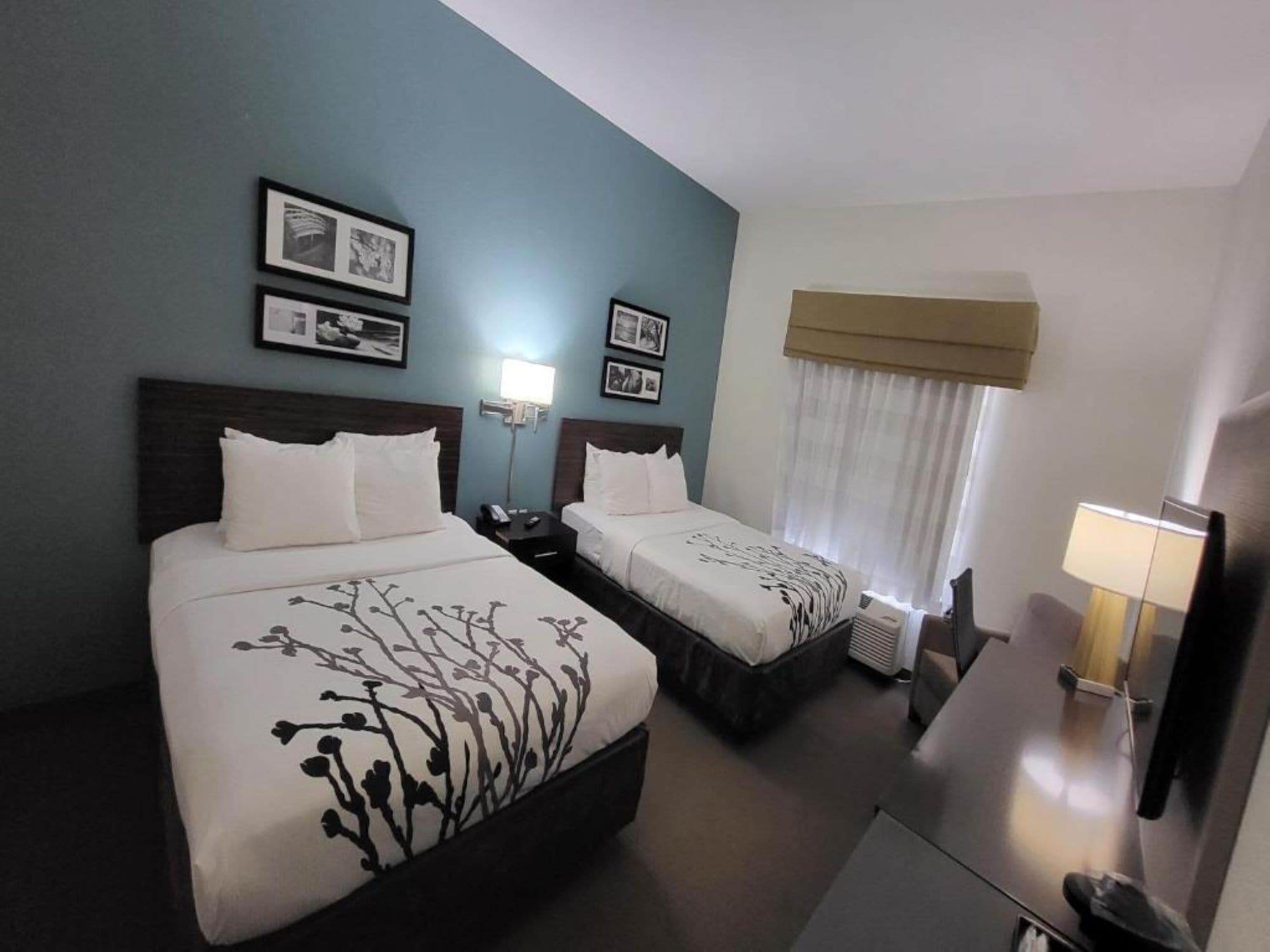 A hotel room with two beds, a desk, a chair, a lamp, and a window with curtains.