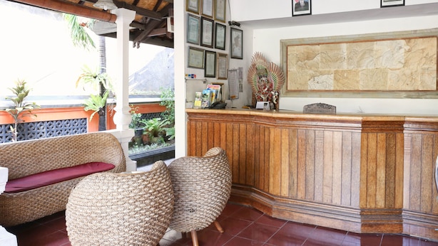 A reception area with a wooden counter, wicker furniture, framed certificates, and a large mural. - RedDoorz @ Pratama Nusa Dua (Nusa Dua)