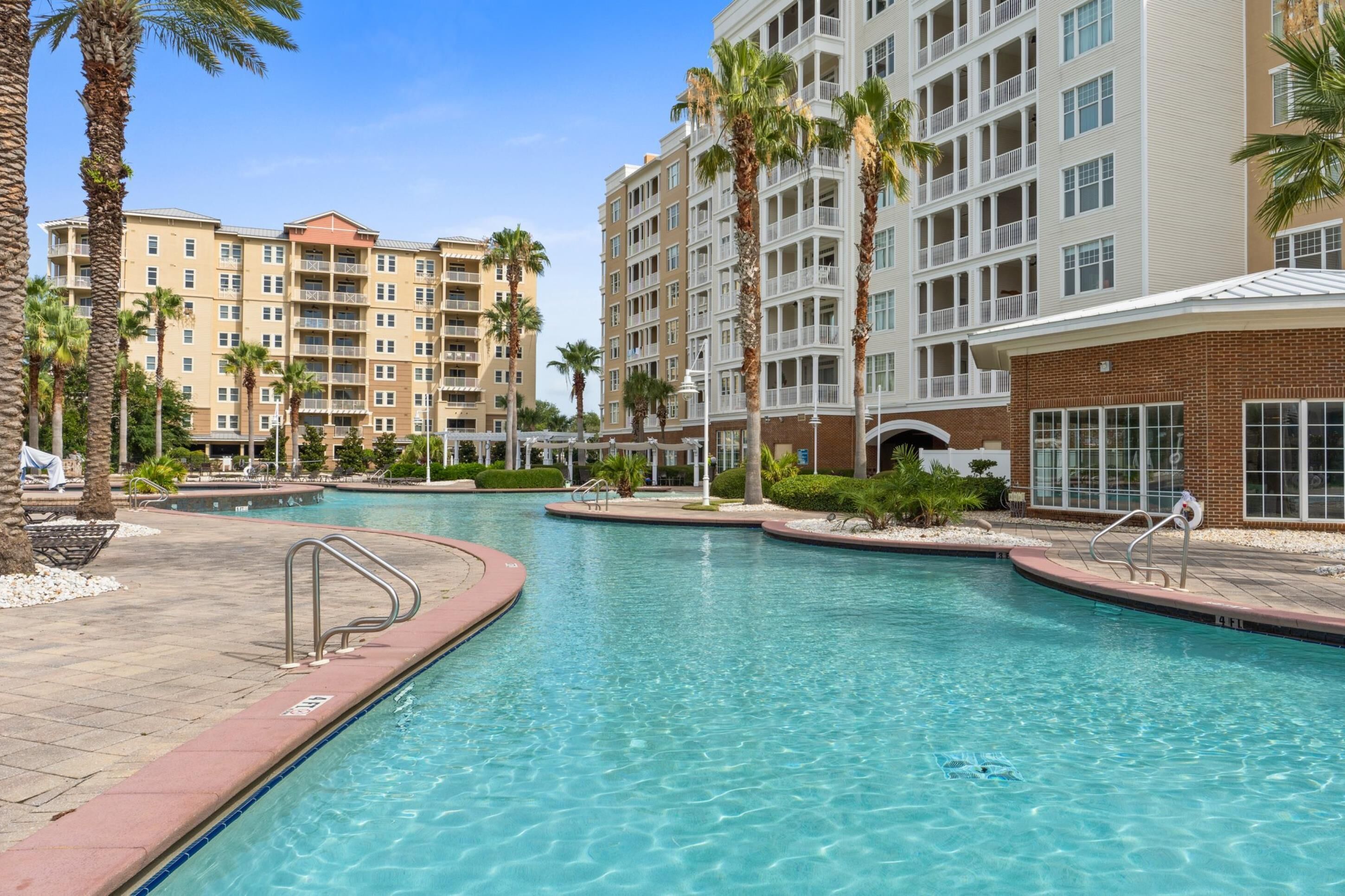 A swimming pool area with a brick building and multiple balconies, surrounded by palm trees.