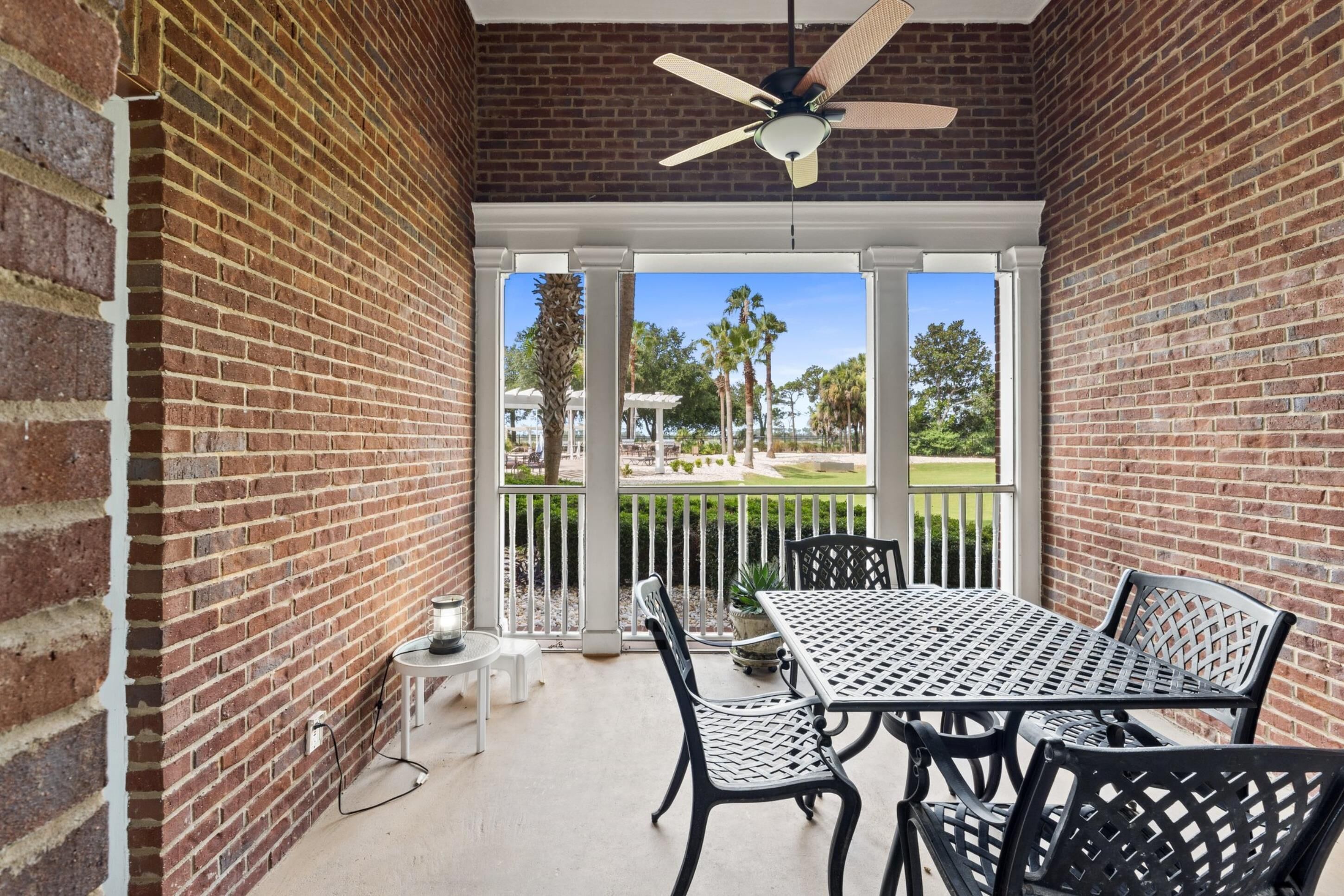 A covered patio with a brick wall, a ceiling fan, a table with chairs, and a view of a landscaped area with palm trees.