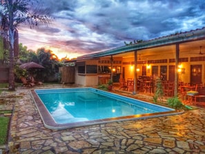 A pool area with stone flooring, a covered seating area with tables and chairs, and a view of the sky at sunset. - Hotel Mariposa (León)