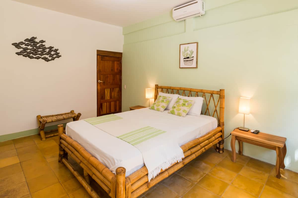 A room with a wooden bed, two bedside tables with lamps, a wooden bench, a door, and a wall decoration.