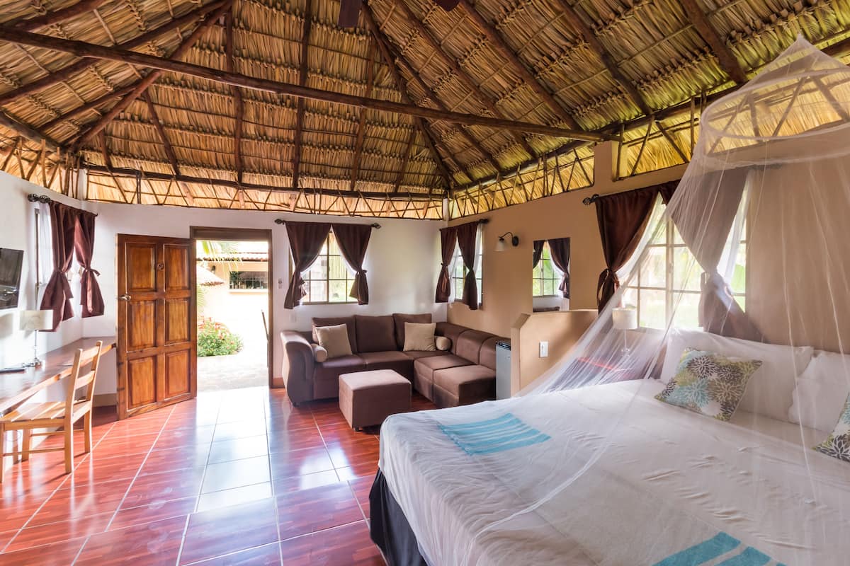 A spacious room with a thatched roof, a large bed with a canopy, a sofa, and a dining area with a table and chairs.
