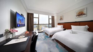 A hotel room with two beds, a desk with a computer, a TV, and a large window with a city view.