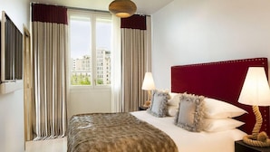 Une chambre d’hôtel avec un grand lit, deux lampes de chevet, une tête de lit rouge et une vue sur les immeubles par la fenêtre.