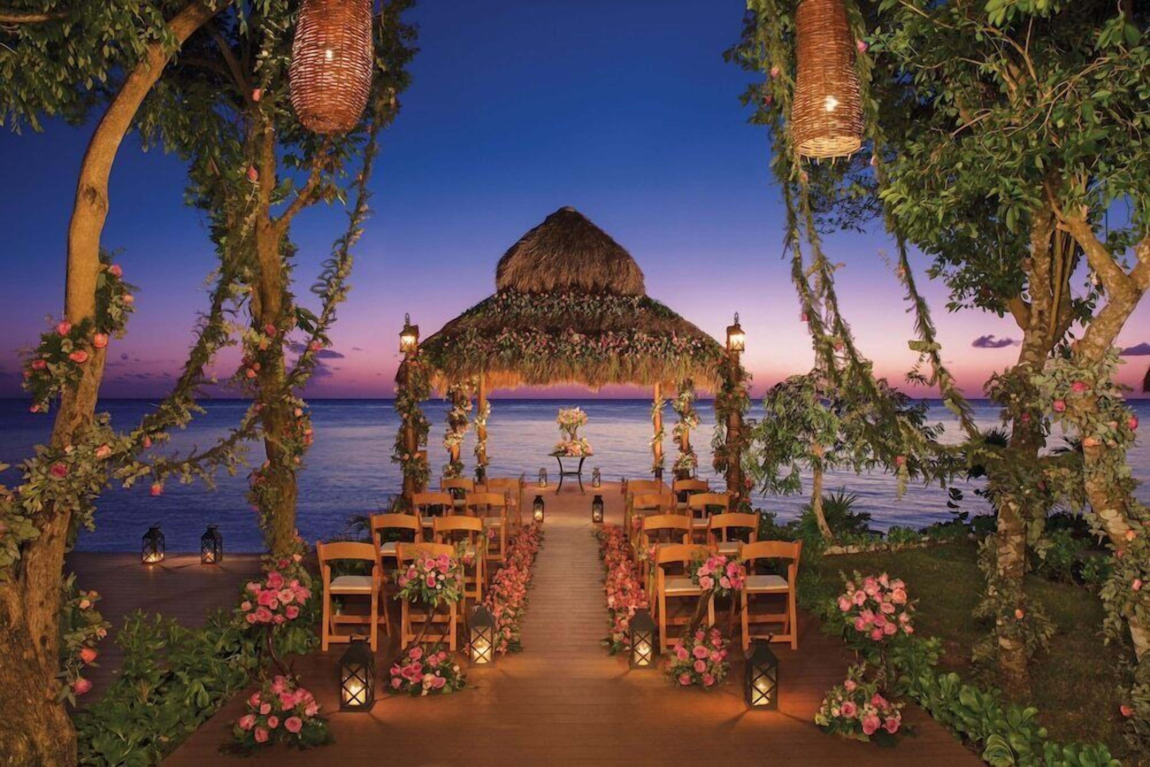 Un lieu de réception en plein air pour les mariages, agrémenté d’une arche en chaume, de chaises en bois, de lanternes et offrant une vue sur la mer.