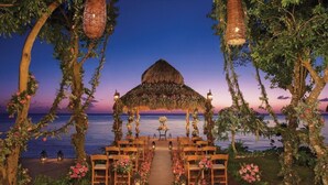 An outdoor wedding venue with a thatched arch, wooden chairs, lanterns, and a view of the sea. - Sabor Cozumel Resort AND SPA (COZUMEL)