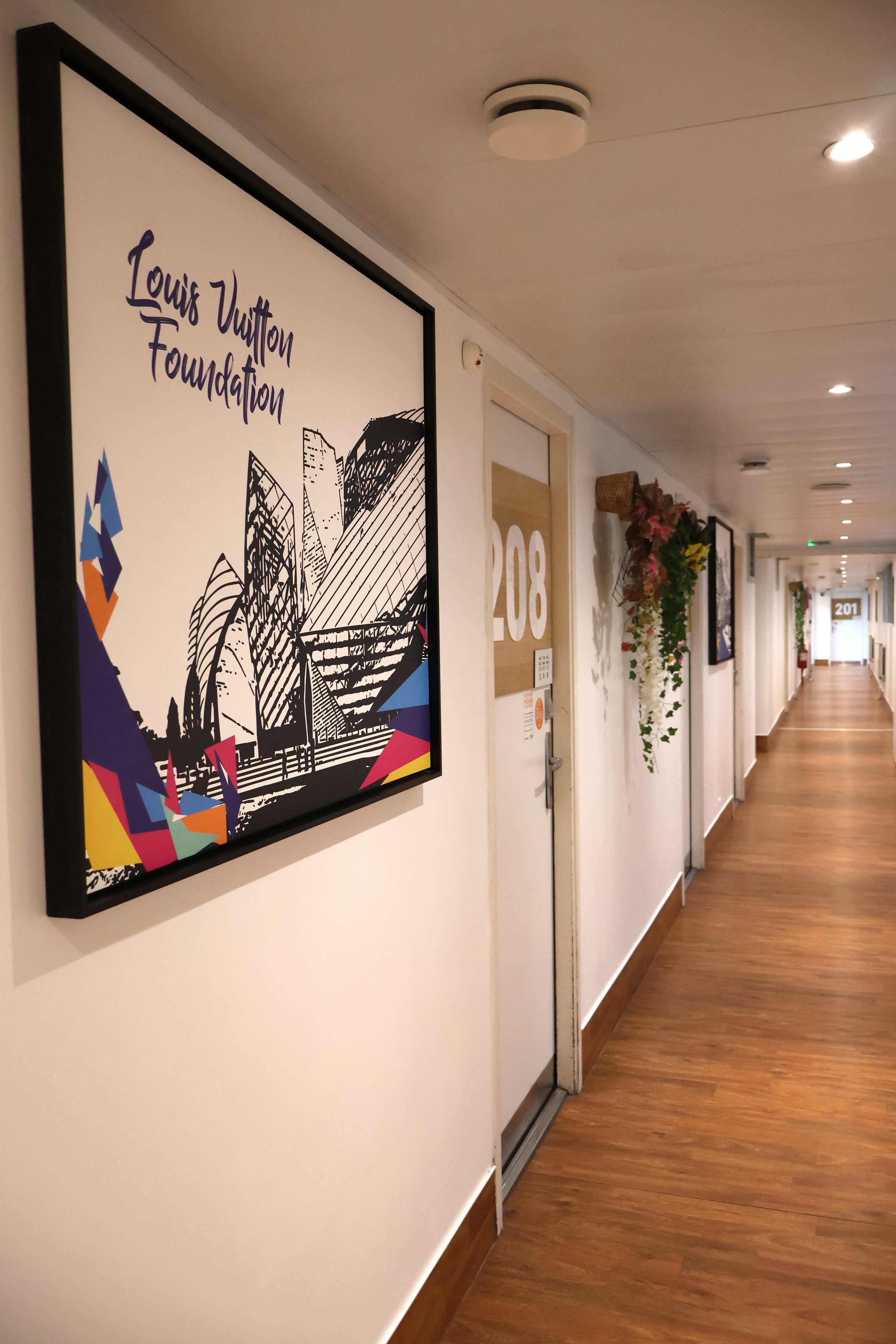 Un couloir orné d’une œuvre d’art encadrée représentant la Fondation Louis Vuitton, une porte portant l’inscription « 208 », et une plante fixée au mur.