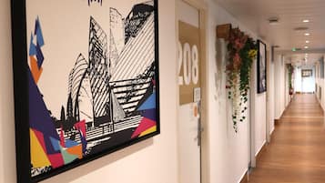 Un couloir orné d’une œuvre d’art encadrée représentant la Fondation Louis Vuitton, une porte portant l’inscription « 208 », et une plante fixée au mur.