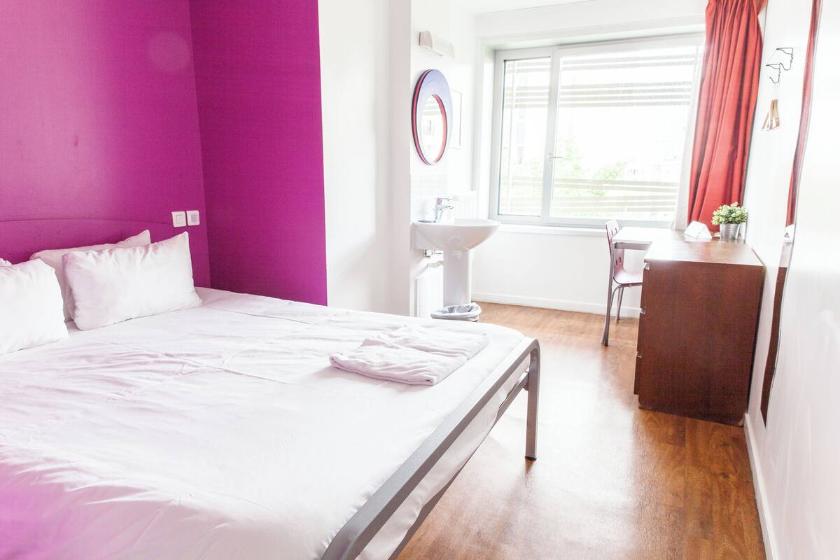 A hotel room with a bed, a desk, a chair, a window with curtains, and a sink.