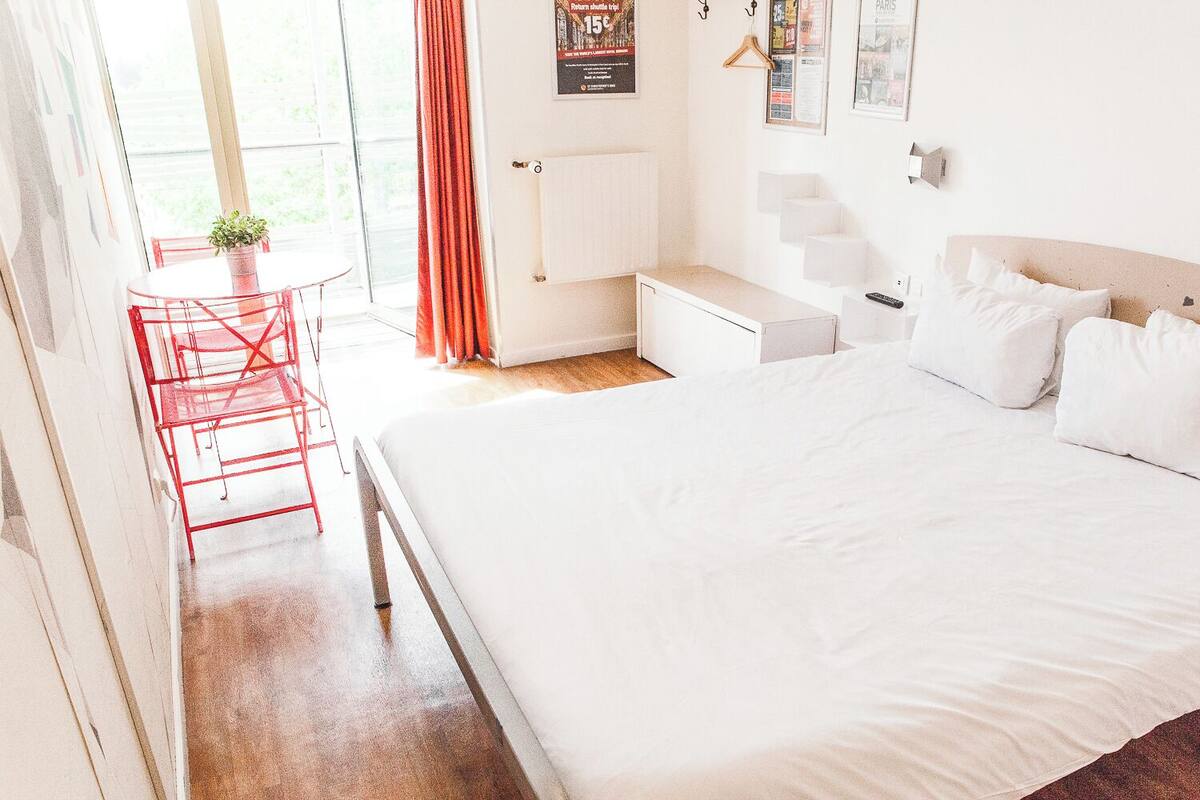 A modern hotel room with a large bed, a small desk, a chair, and a window with curtains.