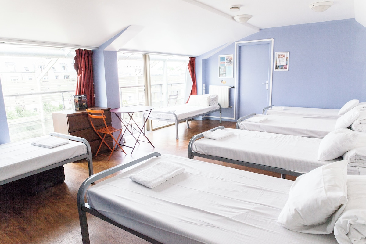 A hotel room with four single beds, a small desk, a chair, and a window with curtains.