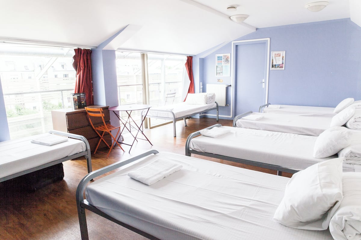 A hotel room with four single beds, a small desk, a chair, and a window with curtains.