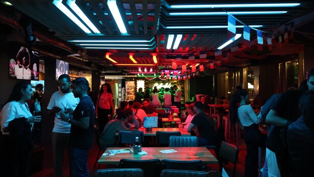 A restaurant with multiple tables and chairs, neon ceiling lights, and a crowd of people. - St. Christophers Paris Canal (Paris)