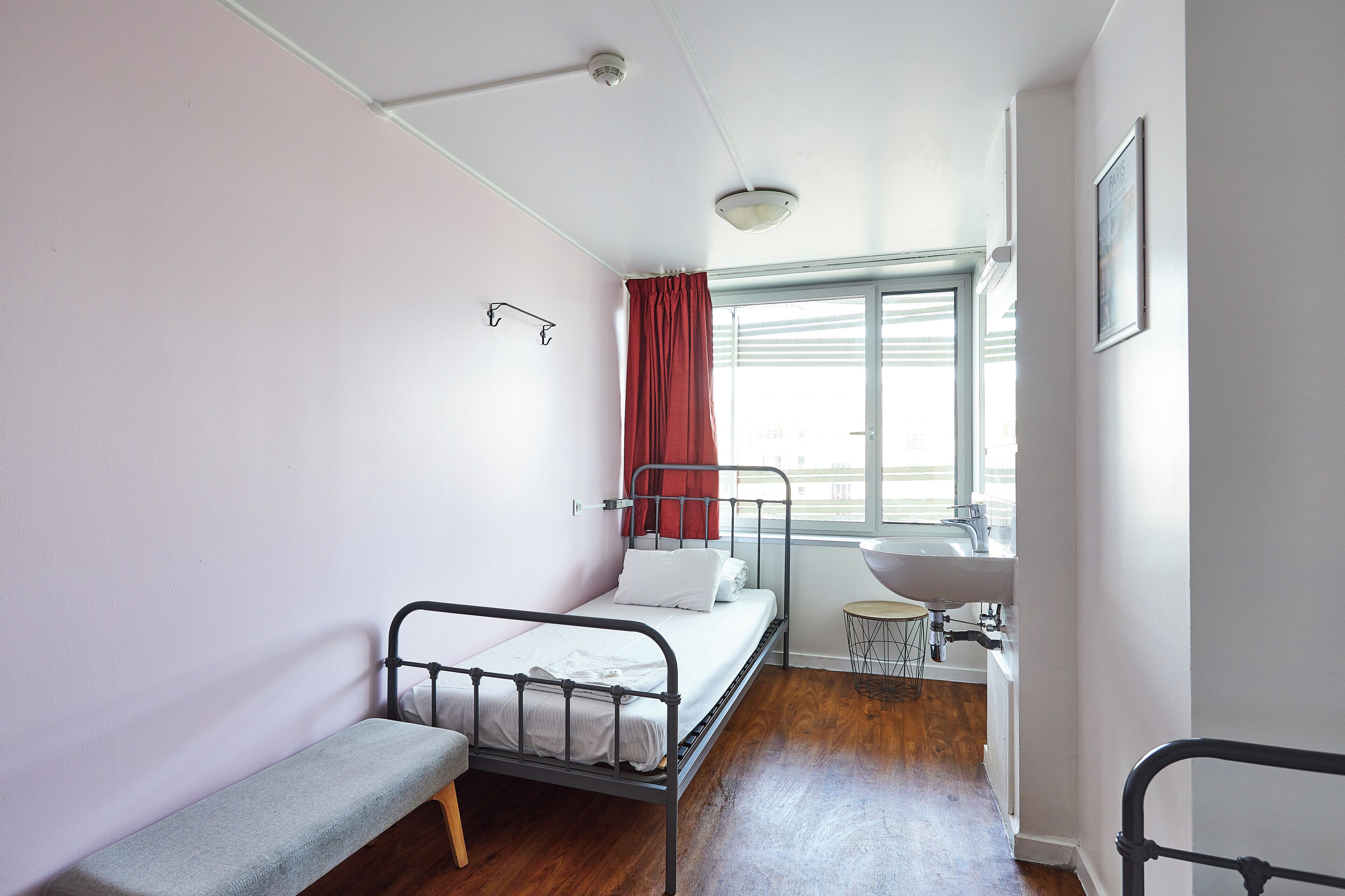 Una habitación pequeña con cama individual, suelo de madera, un banco, una ventana con cortinas rojas, un lavabo y un cuadro en la pared.