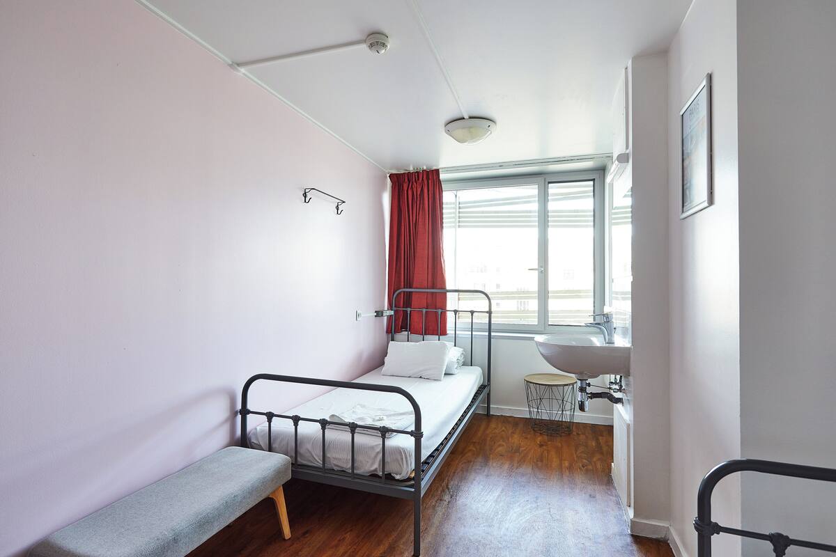 A small, single-bed room with a window, a bedside table, and a built-in sink.