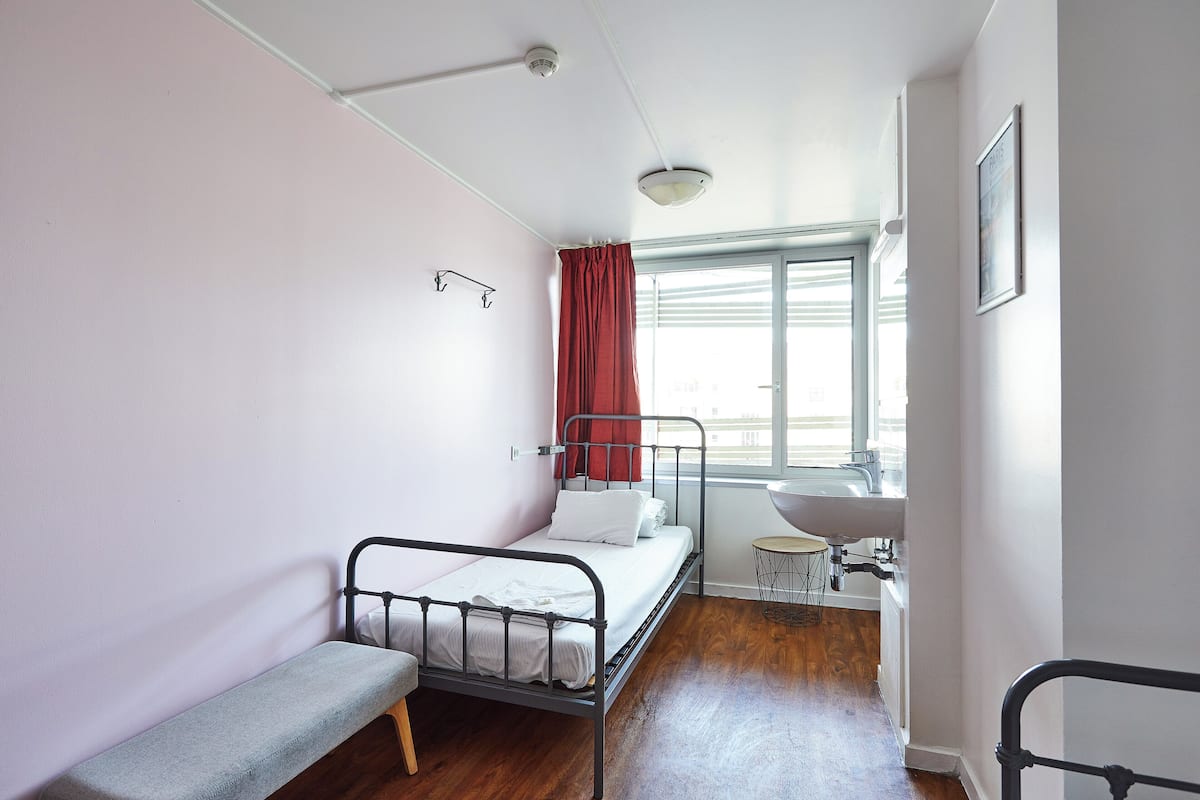 Una habitación pequeña con cama individual, suelo de madera, un banco, una ventana con cortinas rojas, un lavabo y un cuadro en la pared.