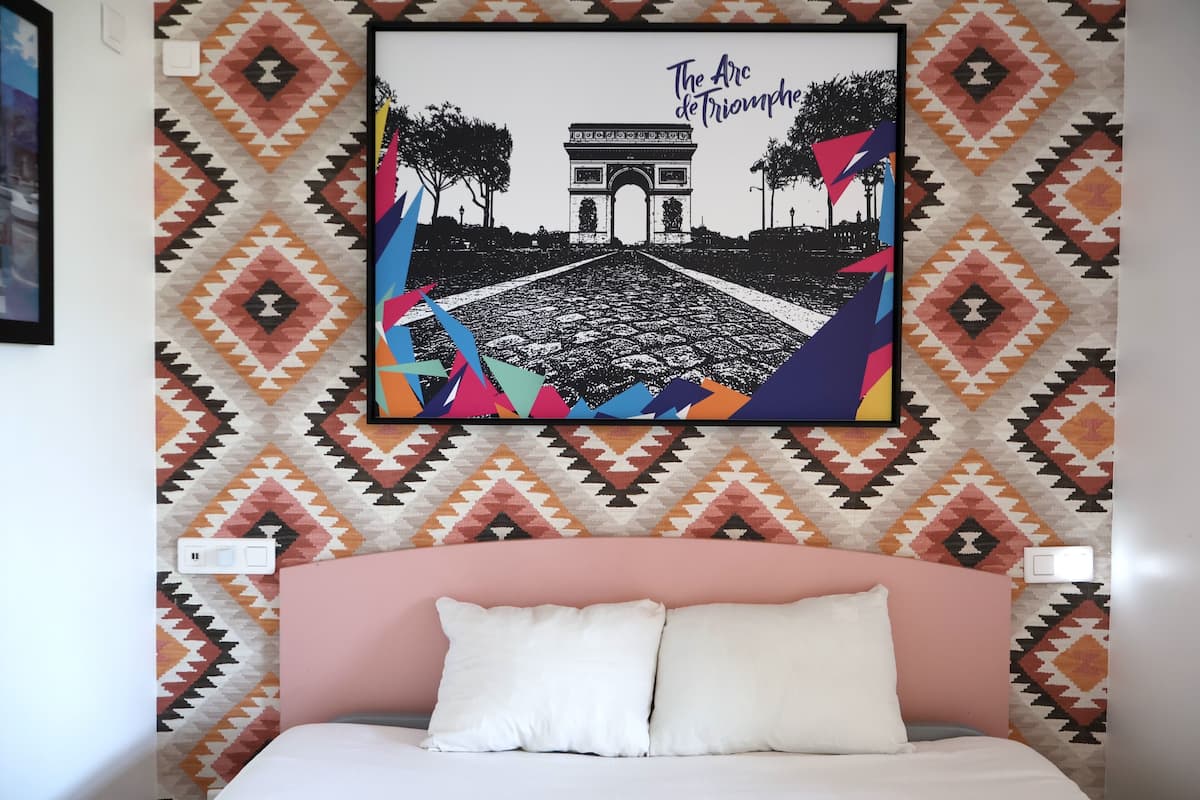 A bed with a pink headboard, a wall with a geometric patterned wallpaper, and a framed picture of the Arc de Triomphe.
