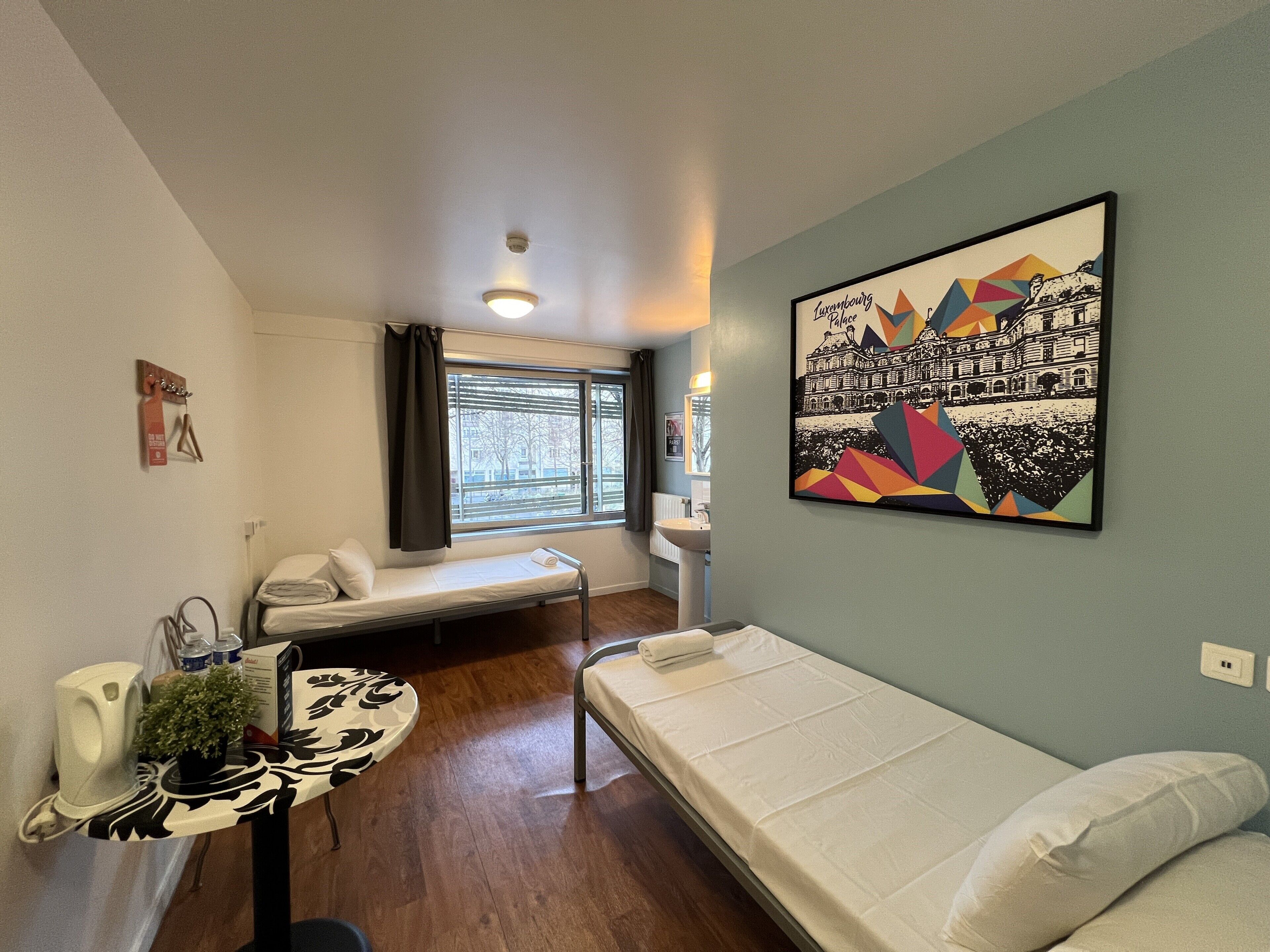 Una habitación pequeña y acogedora con cama, una mesita de noche con planta, ventana con persianas y un cuadro colgado en la pared.