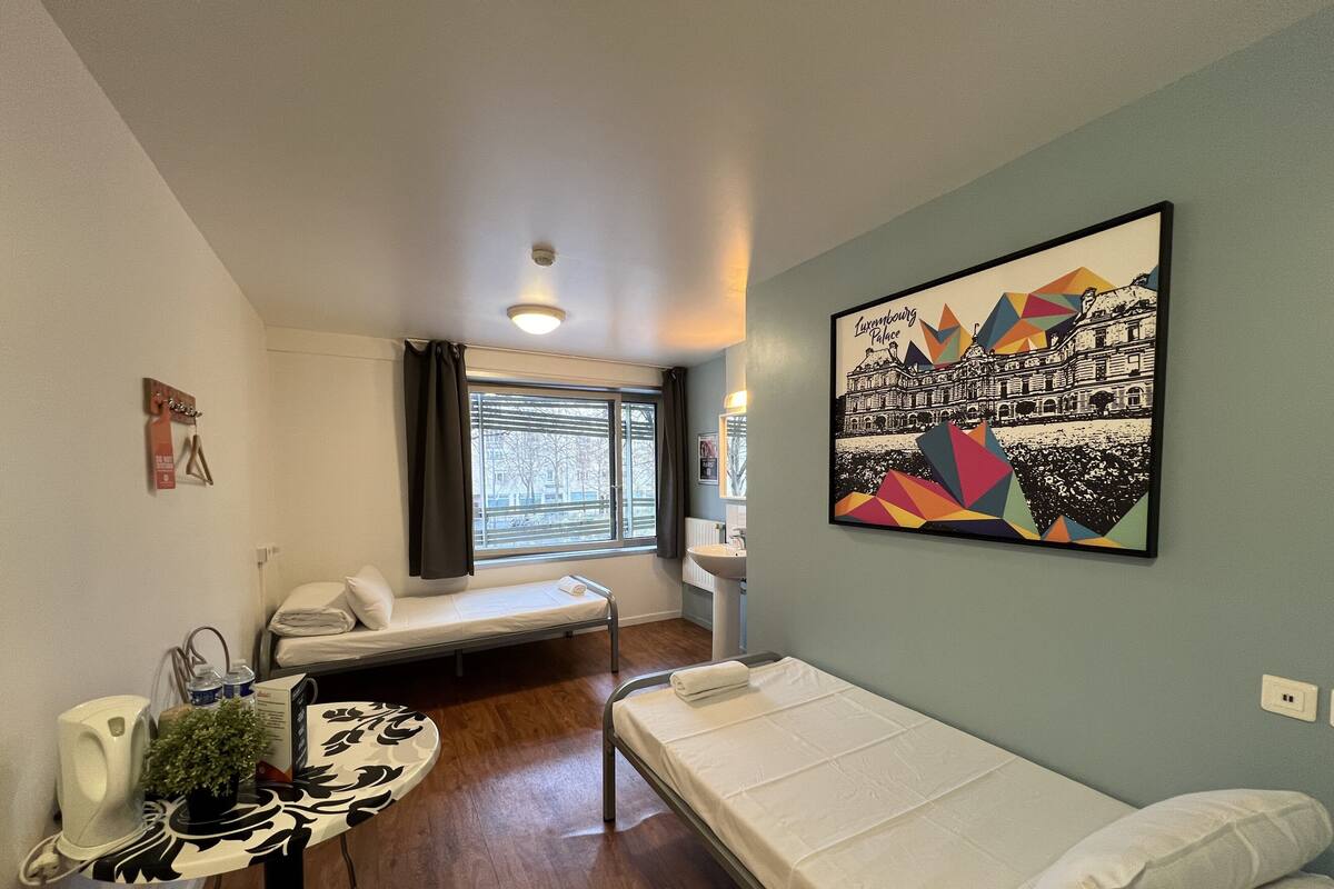 A compact room with a bed, a small table with a plant, a window with blinds, and a wall-mounted picture.