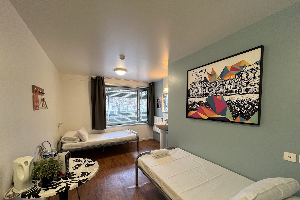A compact room with a bed, a small table with a plant, a window with blinds, and a wall-mounted picture.