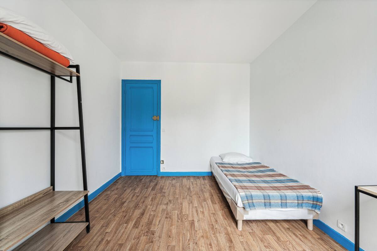 A small, sparsely furnished room with a bunk bed, a single bed, a blue door, and a wooden floor.