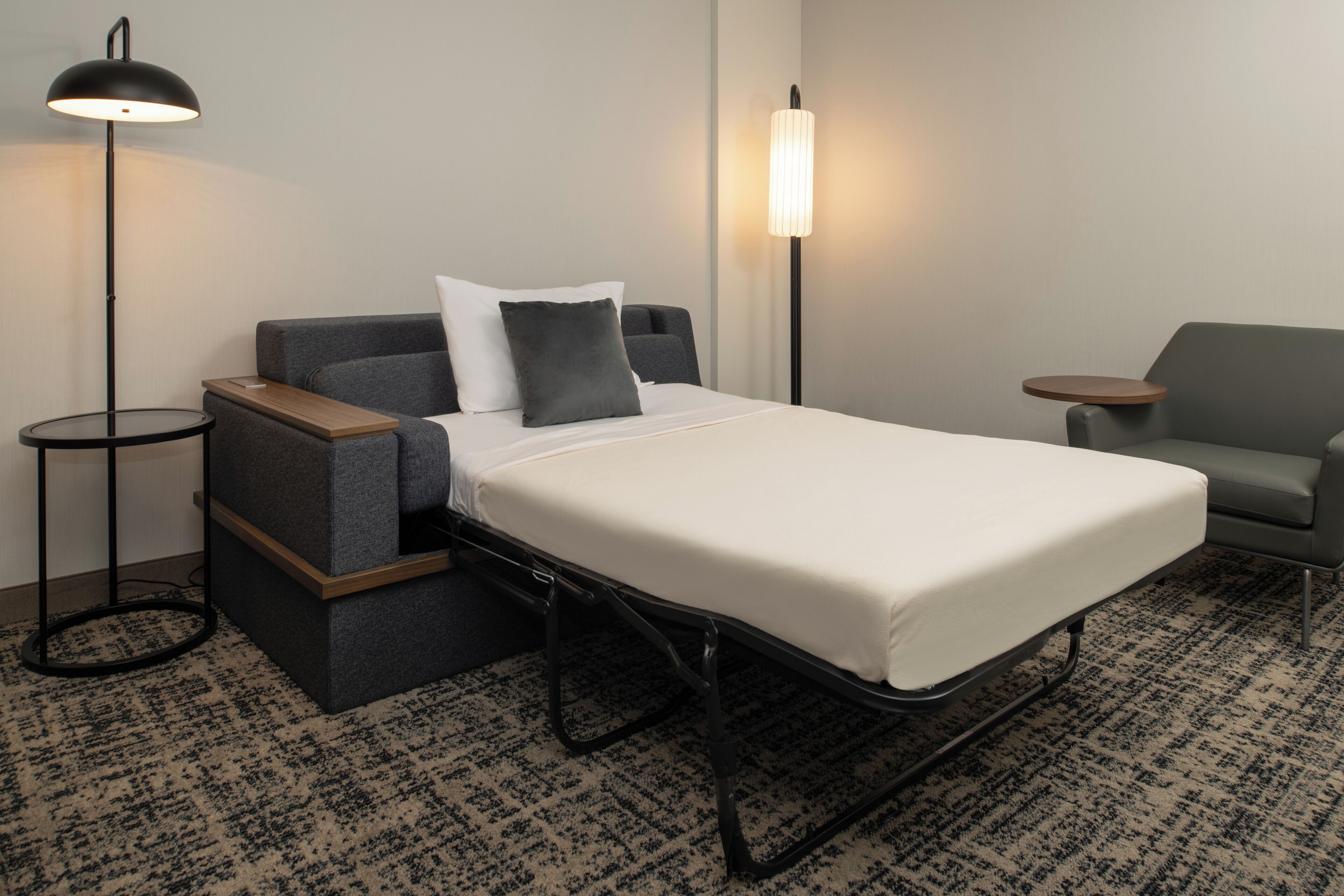 A compact hotel room with a bed, a sofa, a small table, and two floor lamps.