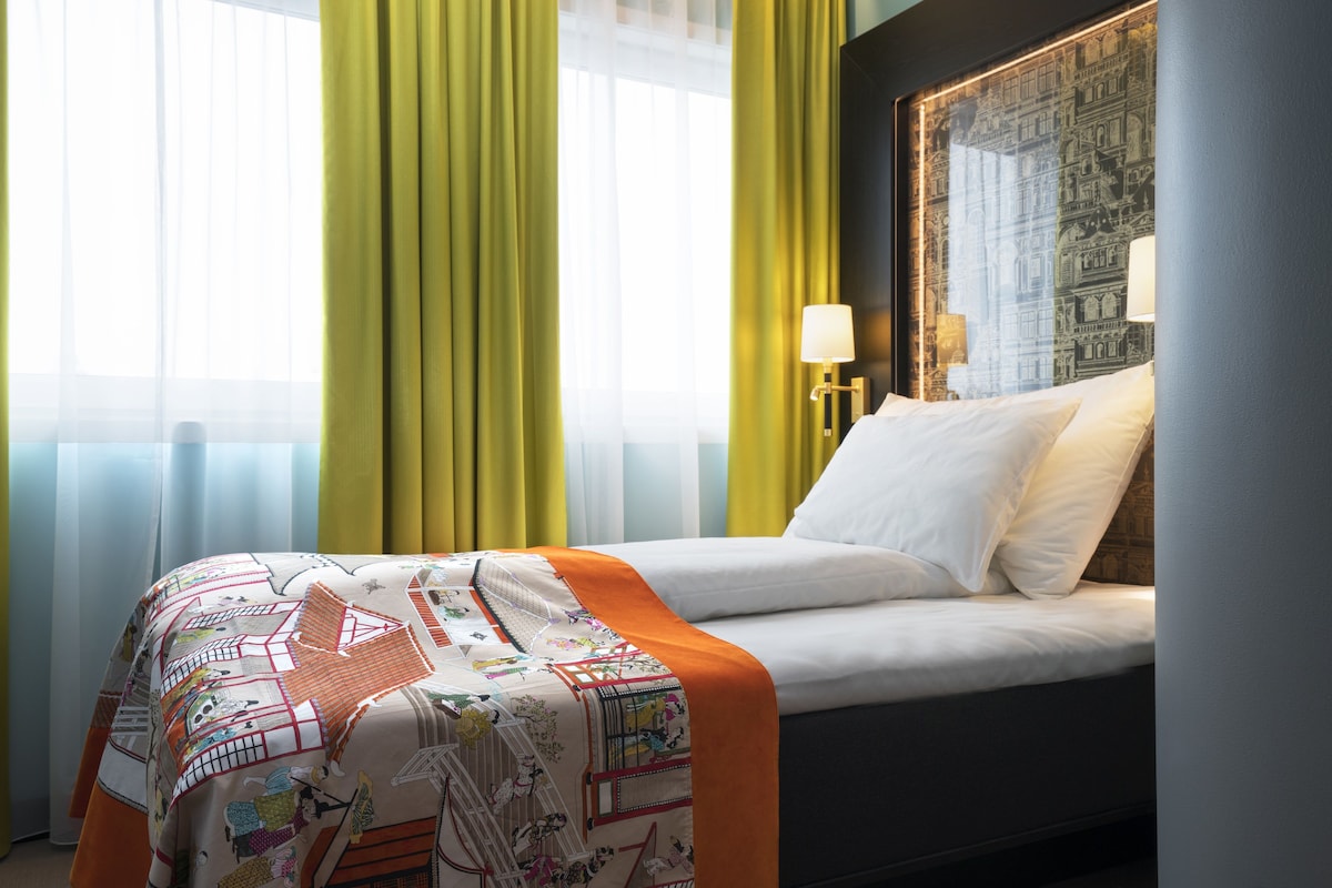 A hotel room with a bed, a window with curtains, a lamp, and a wall with a patterned design.