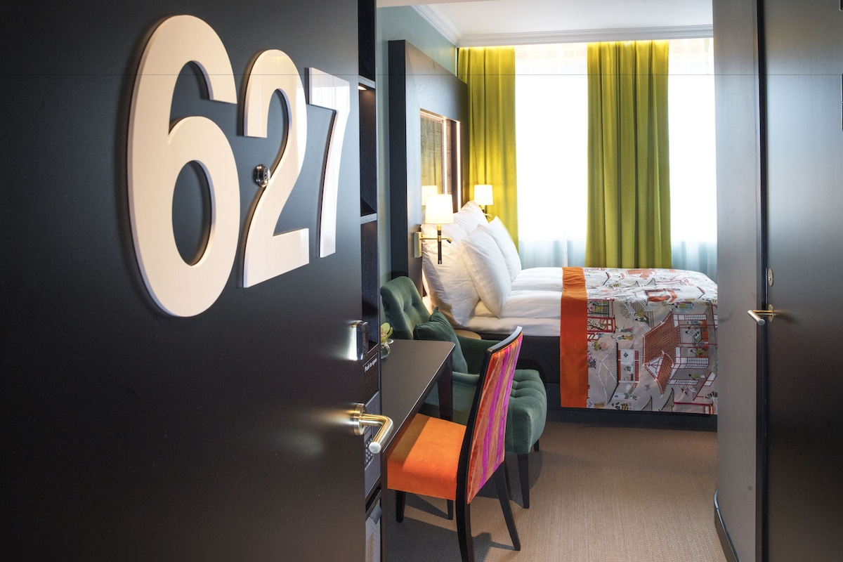 A hotel room with a large number '627' on the wall, a bed with a patterned bedspread, a desk with a chair, and a window with green curtains.