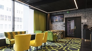 A modern conference room with a large table, yellow and green chairs, patterned carpet, and a city view through large windows.
