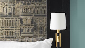 Una cama con un almohadón blanco, una pared con papel tapiz estampado. Se observa una lámpara de pared negra con pantalla blanca y base dorada.